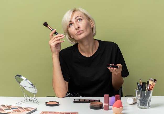 Looking up young beautiful girl sits at table with makeup tools holding powder blush isolated on olive green wall