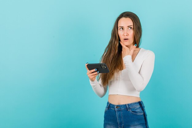 Looking up young girl is thinking by holding smartphone on blue background