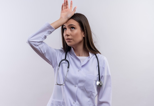 Looking at up young girl wearing stethoscope medical gown put her hand on head on isolated white background