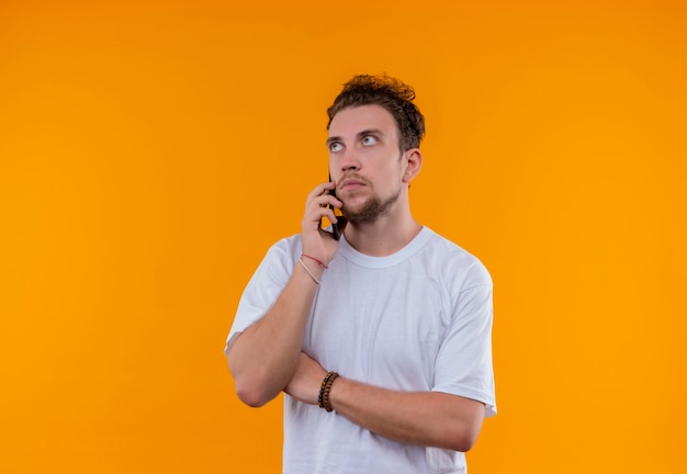 Looking at up young guy wearing white t-shirt speaks on phone crossing hand on isolated orange background