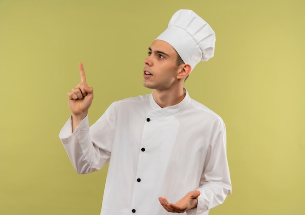 Looking at up young male cook wearing chef uniform points finger to up on isolated green wall with copy space