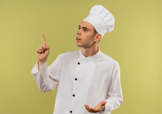 Looking at up young male cook wearing chef uniform points finger to up on isolated green wall with copy space
