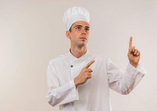 Looking at up young male cook wearing chef uniform points fingers different direction on isolated white wall with copy space