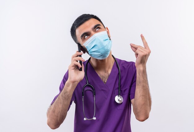 Looking at up young male doctor wearing purple surgeon clothing and stethoscope medical mask speaks on phone on isolated white background