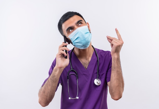 Глядя на молодого врача-мужчину в фиолетовой одежде хирурга и в медицинской маске со стетоскопом, разговаривает по телефону на изолированном белом фоне