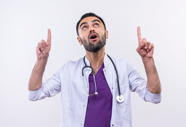 Глядя на молодого врача-мужчину в медицинском халате со стетоскопом, указывает вверх на изолированном белом фоне
