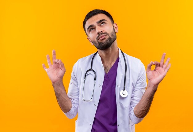 Looking at up young male doctor wearing stethoscope medical gown showing okey gesture on isolated yellow background