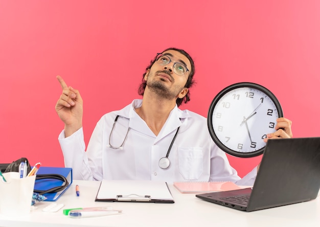 Looking at up young male doctor with medical glasses wearing medical robe with stethoscope sitting at desk