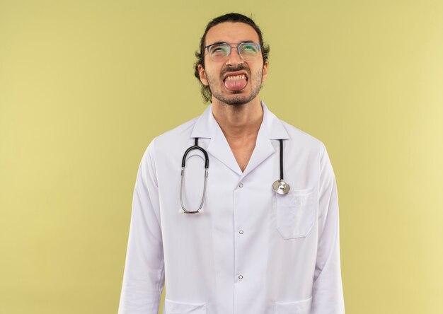 Looking at up young male doctor with optical glasses wearing white robe with stethoscope showing tongue