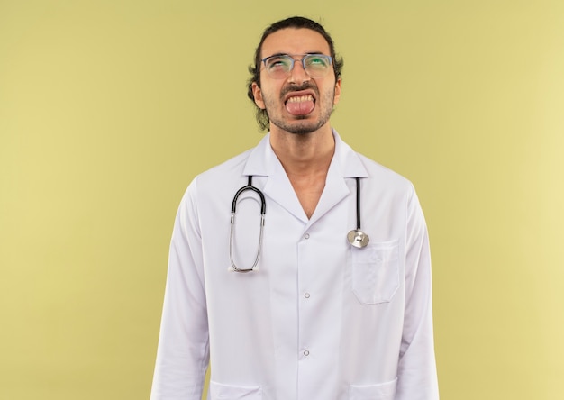 Looking at up young male doctor with optical glasses wearing white robe with stethoscope showing tongue