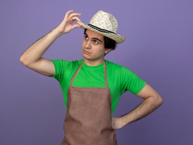 Foto gratuita guardando il giovane giardiniere maschio in uniforme che indossa cappello da giardinaggio tenendo il cappello mettendo la mano sul fianco