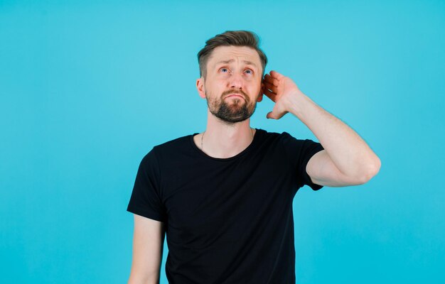 Looking up young man is holding hand on ear on blue background