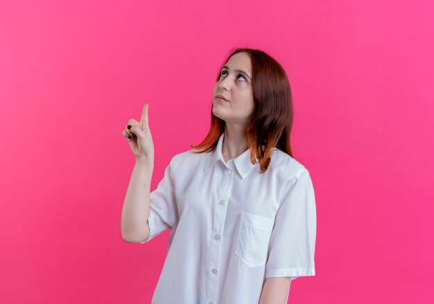 Looking at up young redhead girl points at up isolated on pink background with copy space