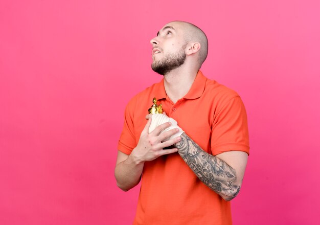 Looking at up young sporty man holding winner cup on chest isolated on pink  wall