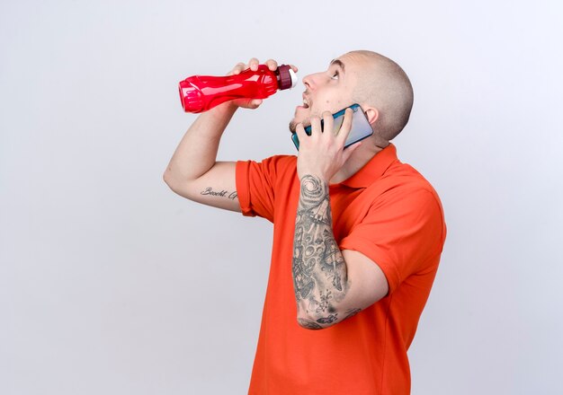 Looking at up young sporty man speakes on phone raising water bottle isolated on white wall