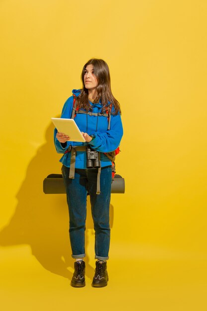 Ищу дорогу с картой. Портрет веселой молодой кавказской туристической девушки с сумкой и биноклем, изолированных на желтом фоне студии. Подготовка к путешествию. Курорт, человеческие эмоции, отдых.