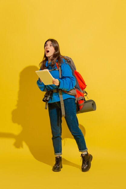 Ищу дорогу с картой. Портрет веселой молодой кавказской туристической девушки с сумкой и биноклем, изолированных на желтом фоне студии. Подготовка к путешествию. Курорт, человеческие эмоции, отдых.