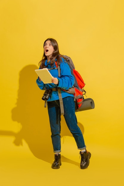 Looking for way with map. Portrait of a cheerful young caucasian tourist girl with bag and binoculars isolated on yellow studio background. Preparing for traveling. Resort, human emotions, vacation.