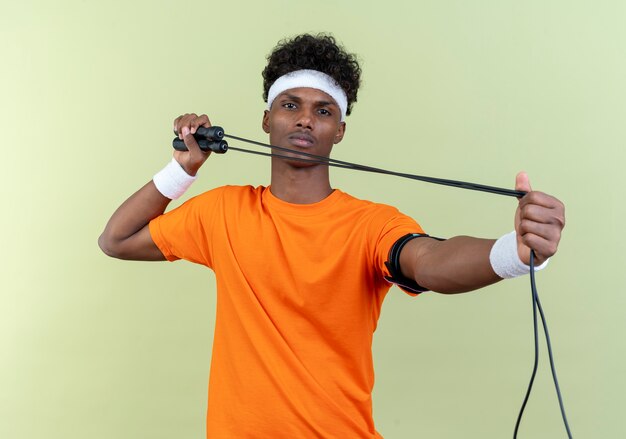 Looking young afro-american sporty man wearing headband and wristband holding jumping rope