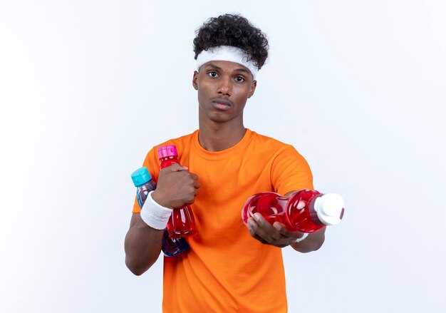 Looking young afro-american sporty man wearing headband and wristband holding out water bottle to camera