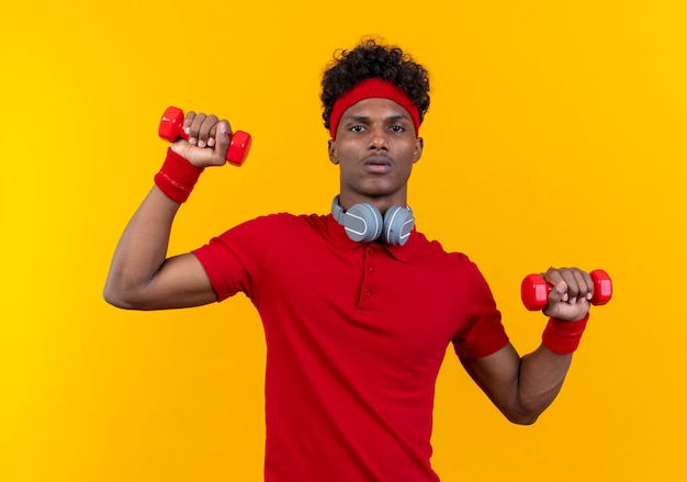 Looking young afro-american sporty man wearing headband and wristband with headphones on neck exercising with dumbbell