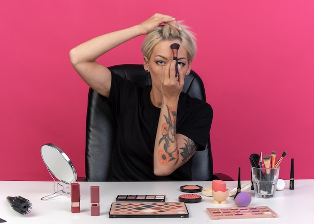 Looking  young beautiful girl sits at table with makeup tools applying powder blush with powder brush on forehead isolated on pink wall