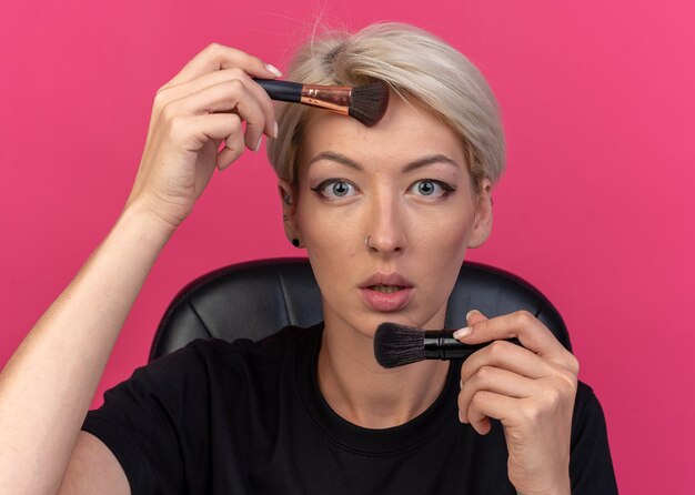 Looking  young beautiful girl sits at table with makeup tools applying powder blush with powder brush isolated on pink wall