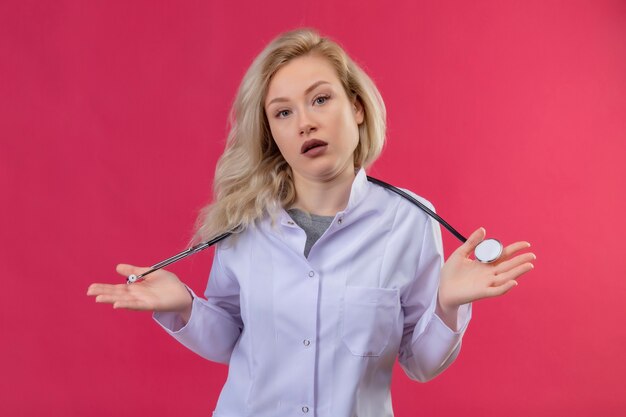 Looking young doctor wearing stethoscope in medical gown soreads hands on red backgroung