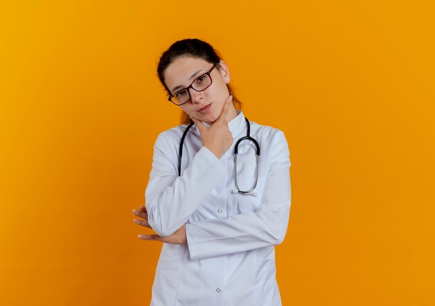 Looking young female doctor wearing medical robe and stethoscope with glasses grabbed chin isolated