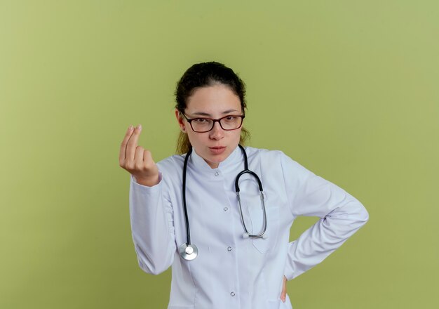 Looking young female doctor wearing medical robe and stethoscope with glasses showing tip gesture putting hand on hip isolated