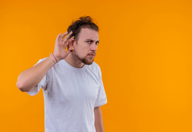 Looking young guy wearing white t-shirt showing listen gesture on isolated orange background