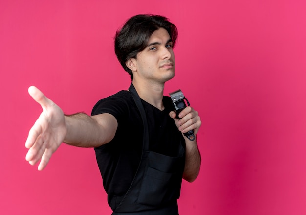 Foto gratuita guardando il giovane barbiere maschio bello in uniforme che tiene le tosatrici e tende la mano