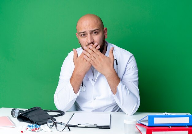 Looking young male doctor wearing medical robe and stethoscope sitting at desk