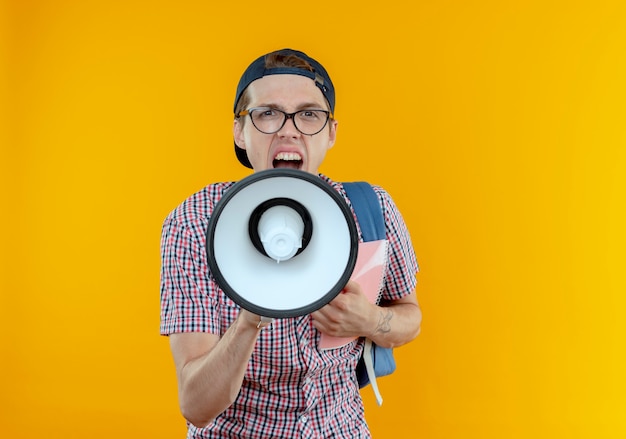 Looking young student boy wearing back bag and glasses and cap speakes on loudspeaker holding notebook