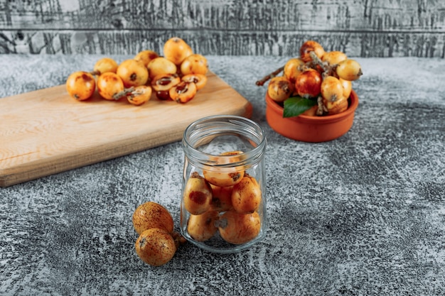 Free photo loquats in a jar, bowl and cutting board on a gray textured background. high angle view.