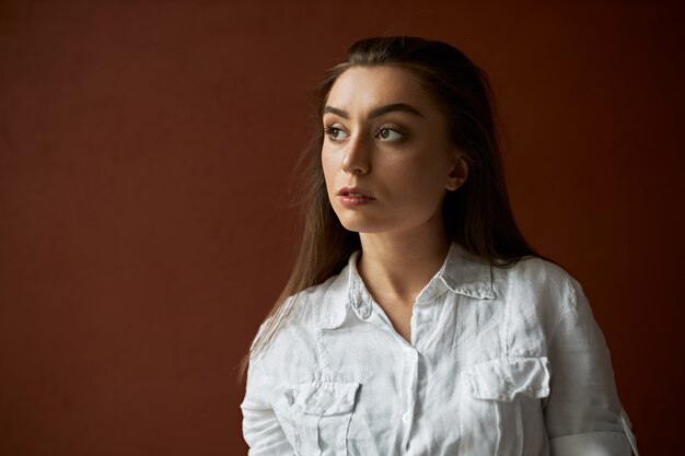 lose up image of serious attractive young woman with brown hair gathered back thinking about something, having pensive deep in thoughts look, posing isolated