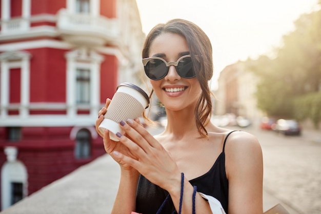 lose up portrait of young beautiful caucasian girl with dark hair in sunglasses and black dress smiling with teeth, drinking coffee, relaxing after long shopping in mall.