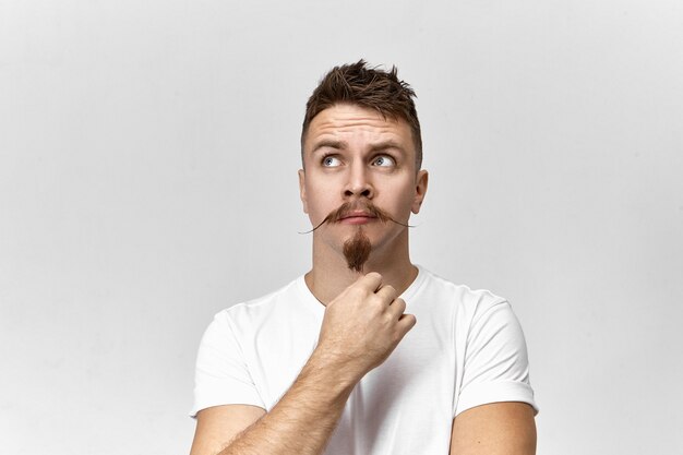 Lost in thoughts. Isolated studio view of attractive stylish young European male hipster with goatee and msutache looking up, having impatient nervous facial expression, waiting for something