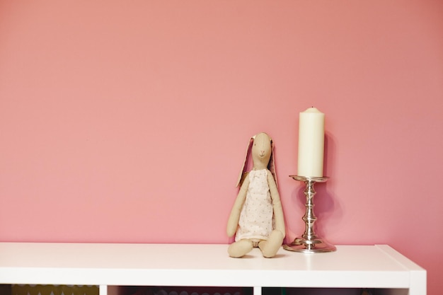 Loth toy rabbit and candle on silver candlestick on white shelf against pink wall