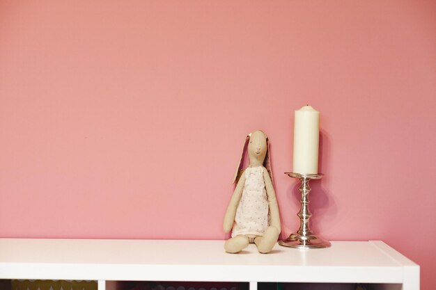 Loth toy rabbit and candle on silver candlestick on white shelf against pink wall