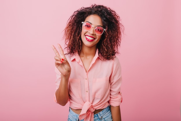 Lovable african girl in cute sunglasses posing with pleasure. Charming curly woman in vintage outfit standing near colorful wall.
