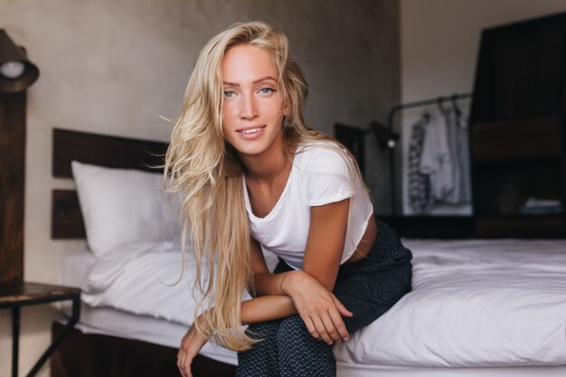 Очаровательная блондинка с голубыми глазами, сидя на кровати. Добродушная женщина в белой футболке проводит утро в своей квартире.