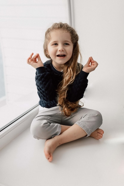 Lovable charming little girl with long wavy hair is sitting on the window and holding up hands with happy charming smile