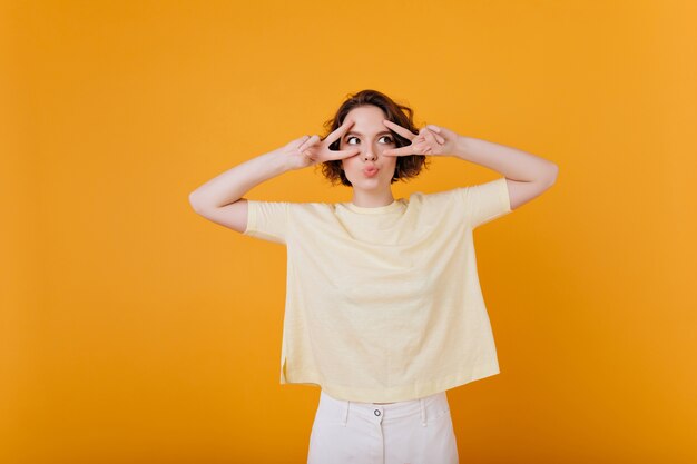 Lovable cute woman with tattoo posing with pleasure on yellow wall. Indoor photo of stylish girl in white pants and oversize t-shirt.