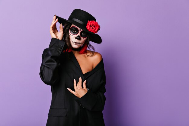 Lovable female model in black outfit posing for halloween photoshoot. Indoor portrait of graceful dark-haired woman with scary face painting.