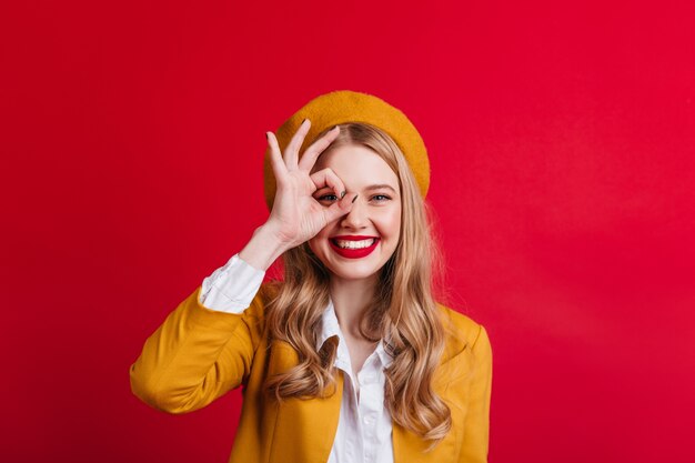 Lovable french girl showing okay sign. Cheerful laughing woman posing on red wall.