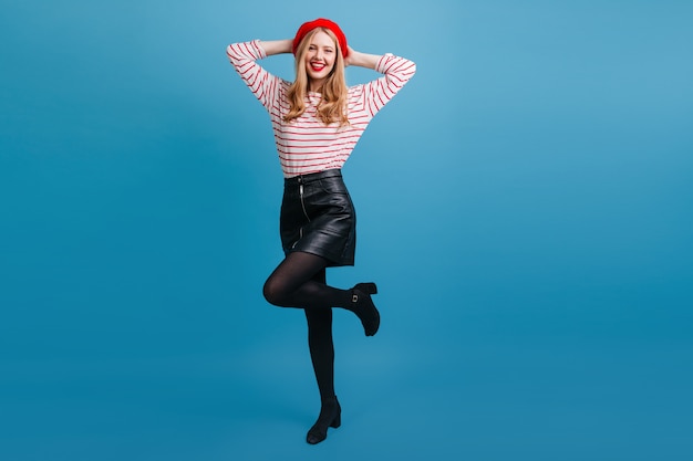 Free photo lovable french girl standing on one leg with smile. carefree blonde young woman dancing on blue wall.