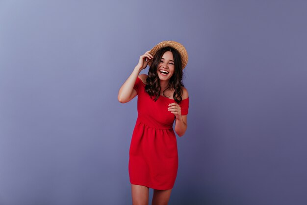 Очаровательная девушка с каштановыми волосами смеется на фиолетовой стене. Фотография в помещении радостной брюнетки в красном платье и летней шляпе.