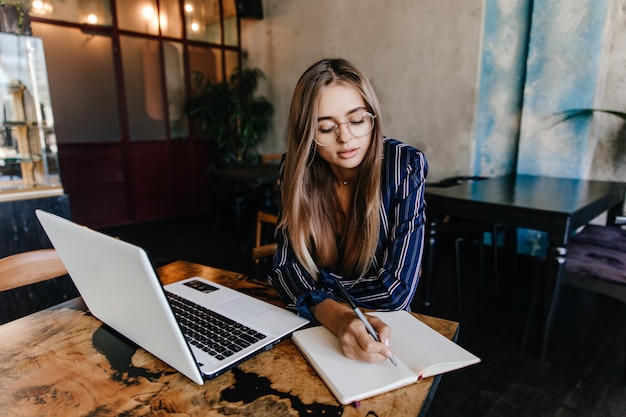 Free photo lovable girl writing something in her notebook. indoor shot of adorable long-haired woman using white laptop in cafe.