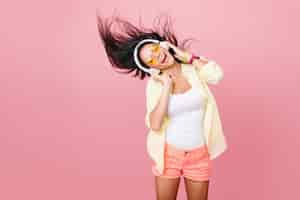 Free photo lovable latin woman in white tank-top dancing with good music and waving hair. indoor portrait of graceful active asian girl in pink shorts relaxing in headphones.